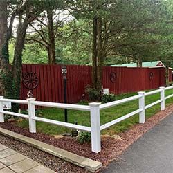 White fence bordering a yard with a red wooden fence, wagon wheels, and lanterns mounted on a grassy area, surrounded by trees.