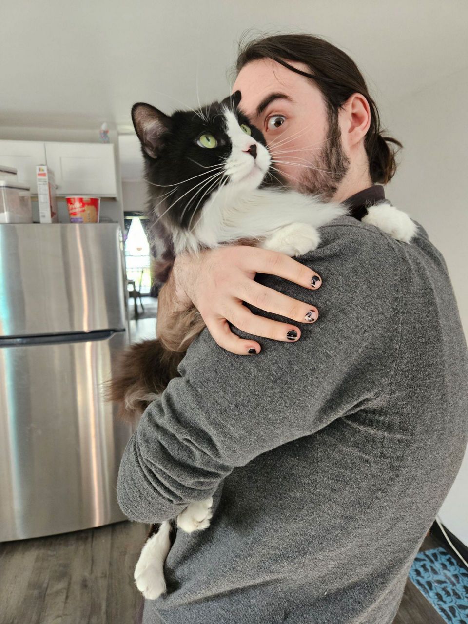Gavin -Daycare & Animal Care Attendant holds a black and white cat close, standing in a kitchen environment with a visible fridge in the background.