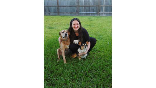 Kings Crossing Kori Piacenti – Assistant Manager in a grassy yard, smiling between two dogs: a brown, medium-sized dog and a tricolor corgi.
