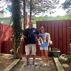 A couple standing on a garden path holding three small dogs, surrounded by a red wooden fence and lush greenery.