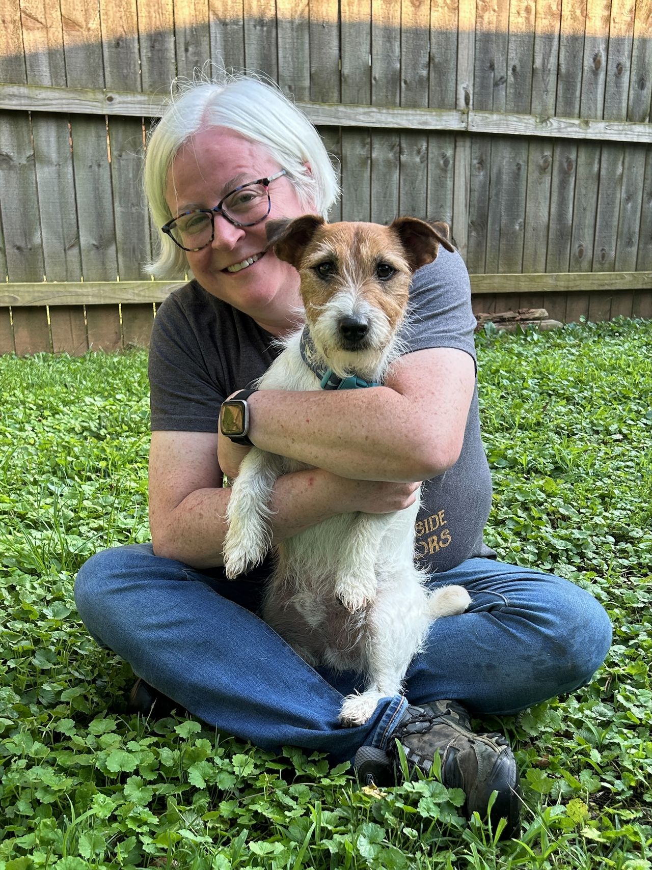A center trainer on grass in a backyard, smiling and holding a small brown and white dog close, with a wooden fence in the background.