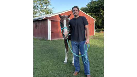 Dr. Braxton Norris -Associate Veterinarian stands beside a brown and white horse with a blue halter in front of a red barn.