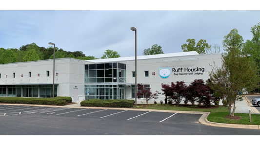 Modern building with large windows, labeled "Ruff Housing Dog Daycare and Lodging," surrounded by trees and greenery, with an empty parking lot.
