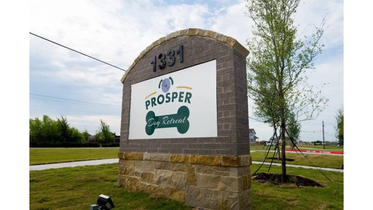 Stone sign for "Prosper Dog Retreat" with dogbone design, surrounded by grassy area, trees, and a cloudy sky in the background.