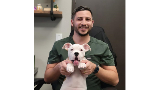 Dr. Brandon Balsino -Veterinarian smiling while holding a happy white puppy by its front paws in an office setting.