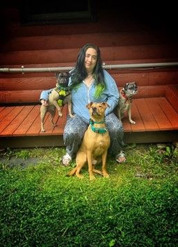 Jessica -Daycare & Animal Care Attendant sits on wooden steps with three dogs; two pugs wearing harnesses and a larger dog sitting in front.