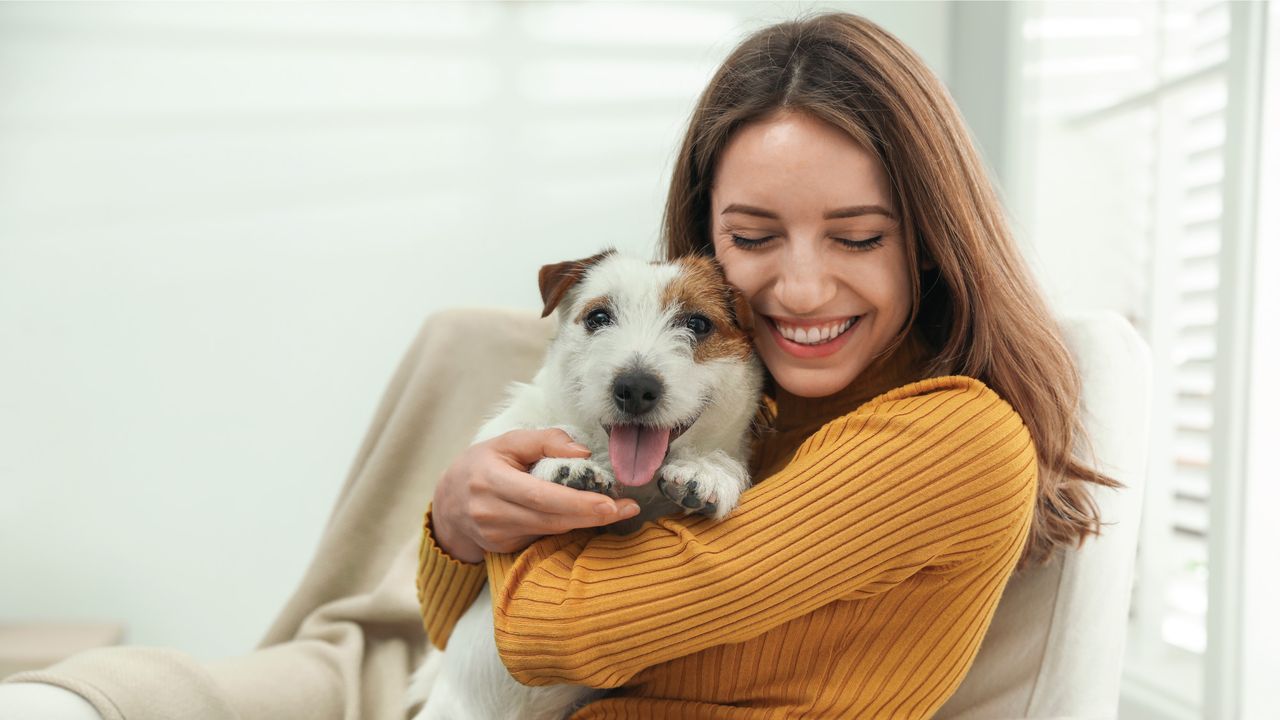 A woman in a mustard sweater happily hugs a small, fluffy dog while sitting indoors. Both are smiling and appear content.