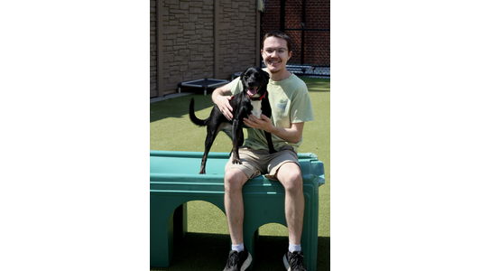Ruff Housing Witt Street Staff Member Jakob sitting on the green plastic play structure, gently holding a happy black and white dog that has its front paws resting on his lap.