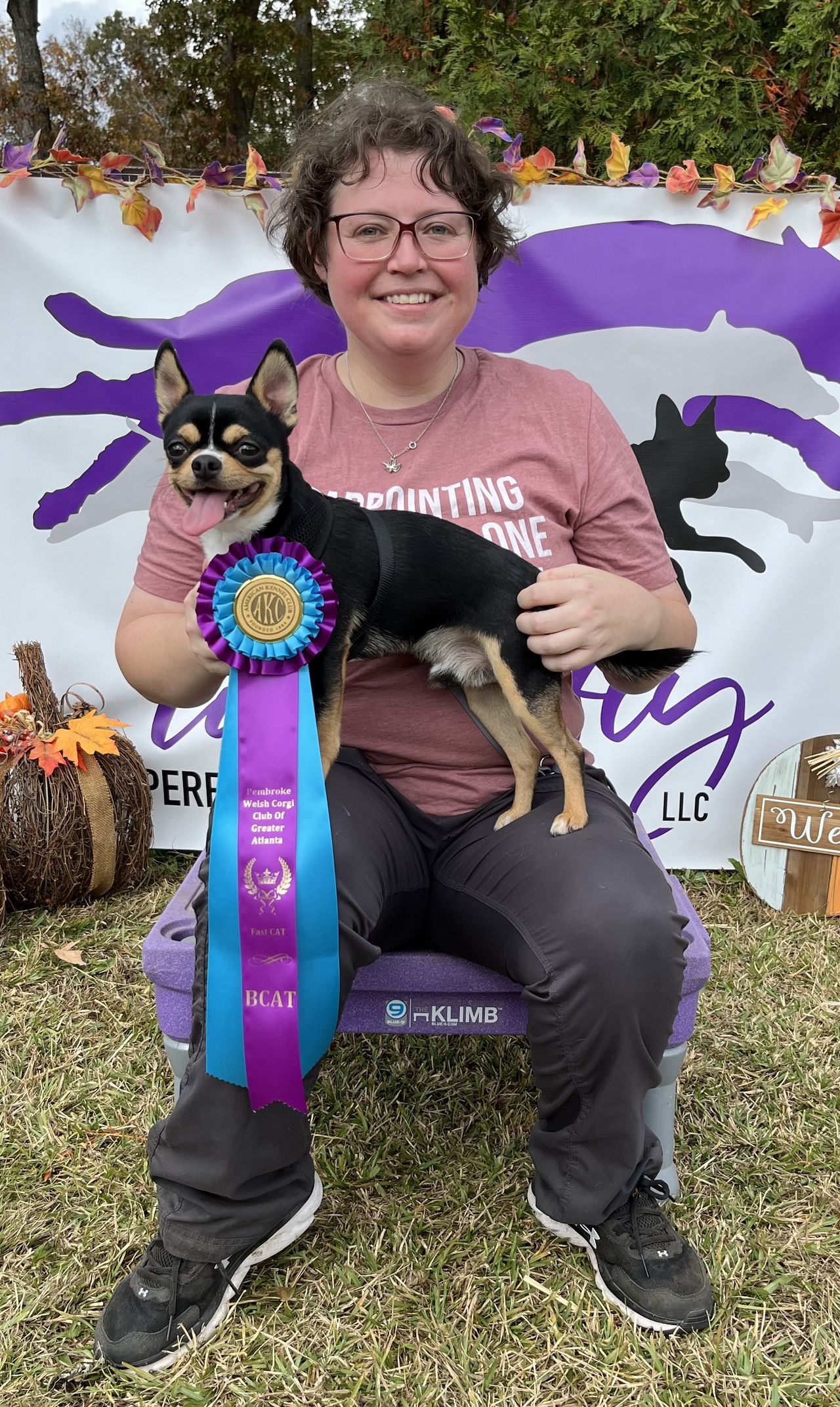 Trainer outdoors holding a small dog with a colorful award ribbon, smiling in front of a decorated background.