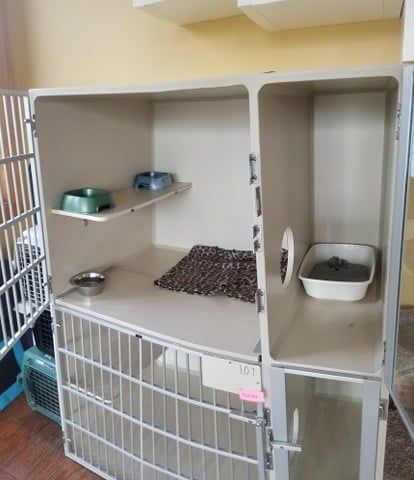 A cat kennel with food and water bowls, a soft blanket, a litter box, and an attached compartment, all enclosed by metal bars.