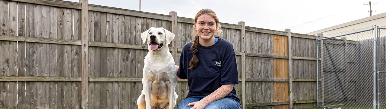 A female staff member sitting on a bench next to a happy white dog, both in a fenced yard.