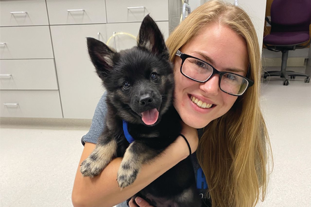 A woman with glasses smiling and holding a fluffy black and tan puppy in a room with white cabinets.