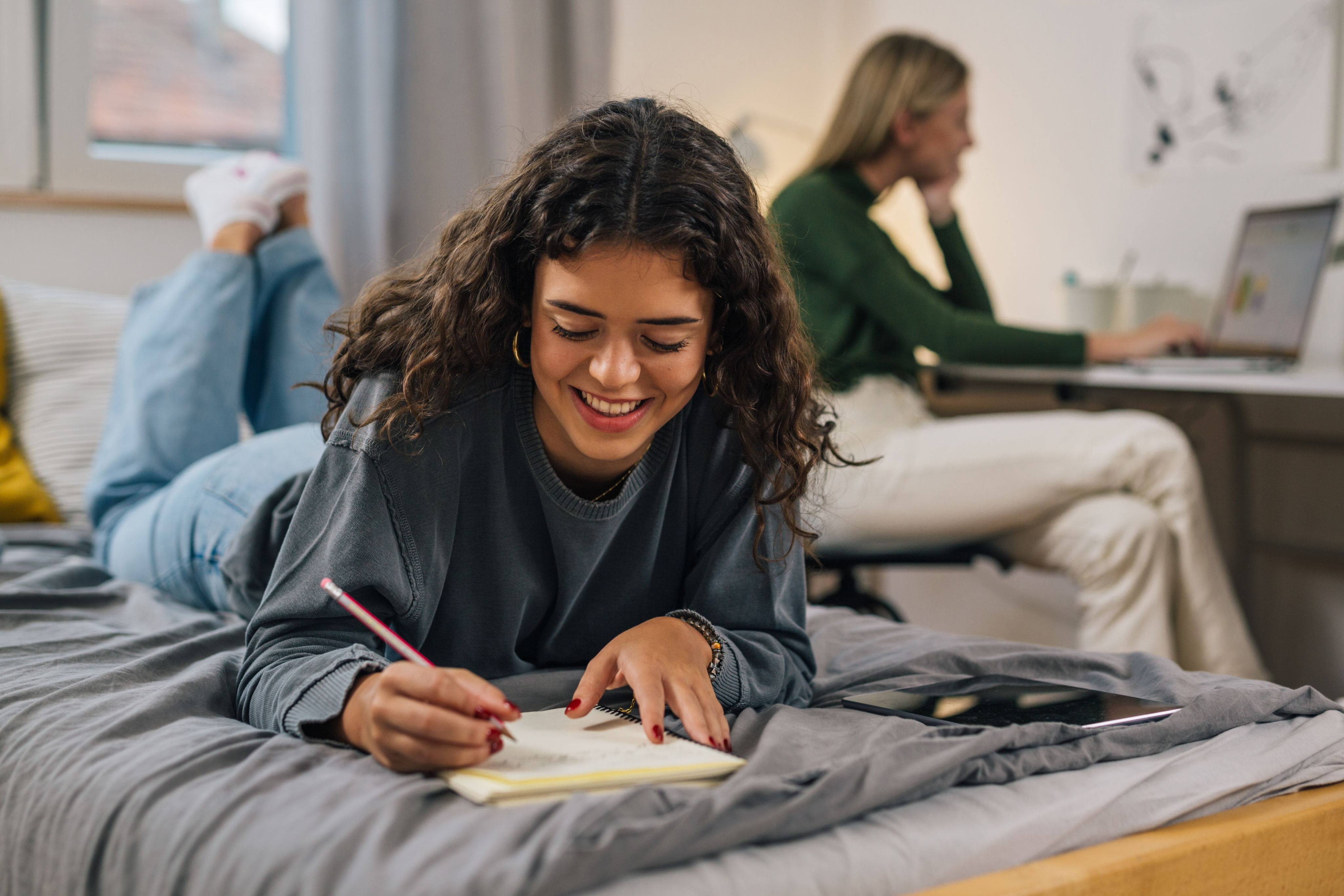Student writing in a notebook on her dorm bed while a roommate studies at a desk in the background