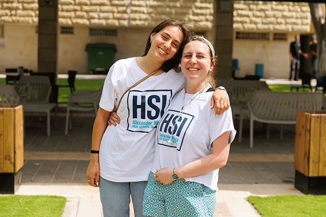 Two smiling students in HSI t-shirts with their arms around each other on campus