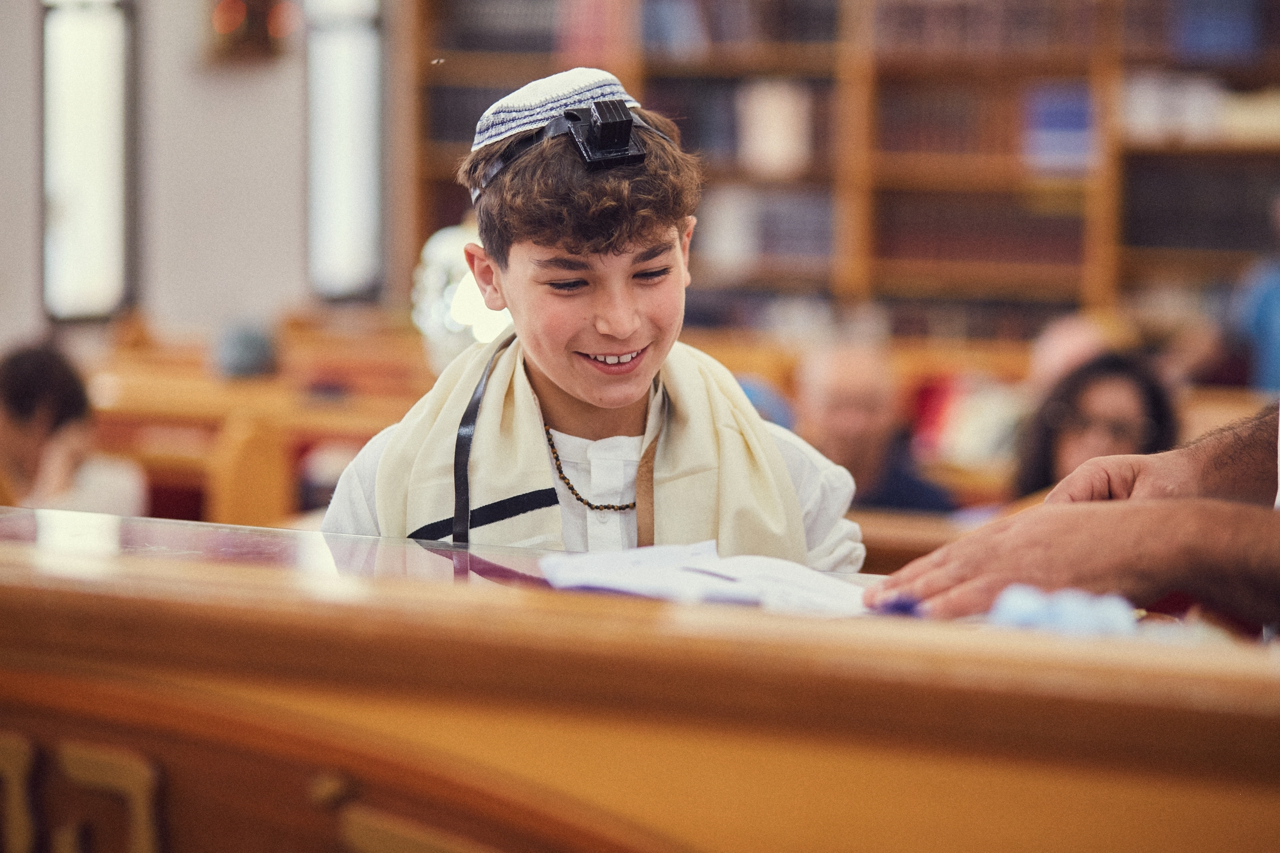 A boy wearing a kippah, tallit, and tefillin smiling at a synagogue