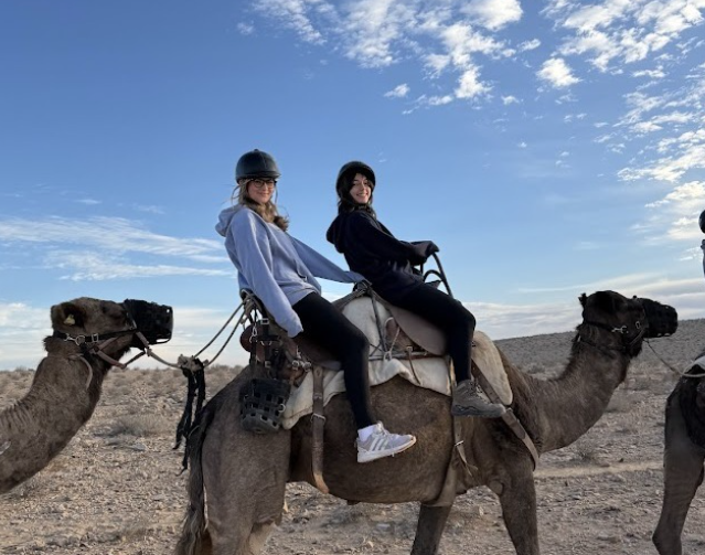 Two students riding camels in the desert