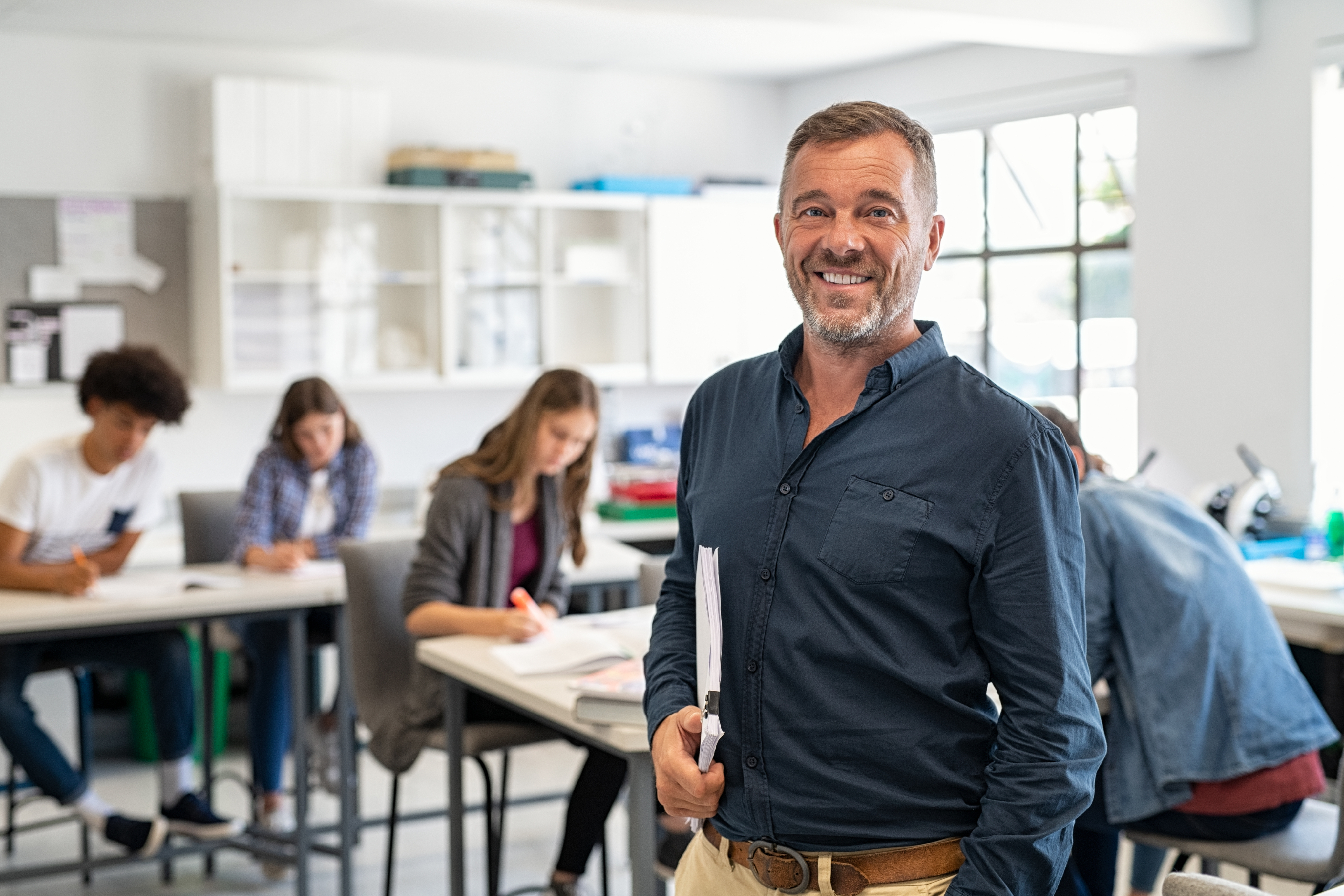 Teacher smiling in a classroom with students studying at desks behind him