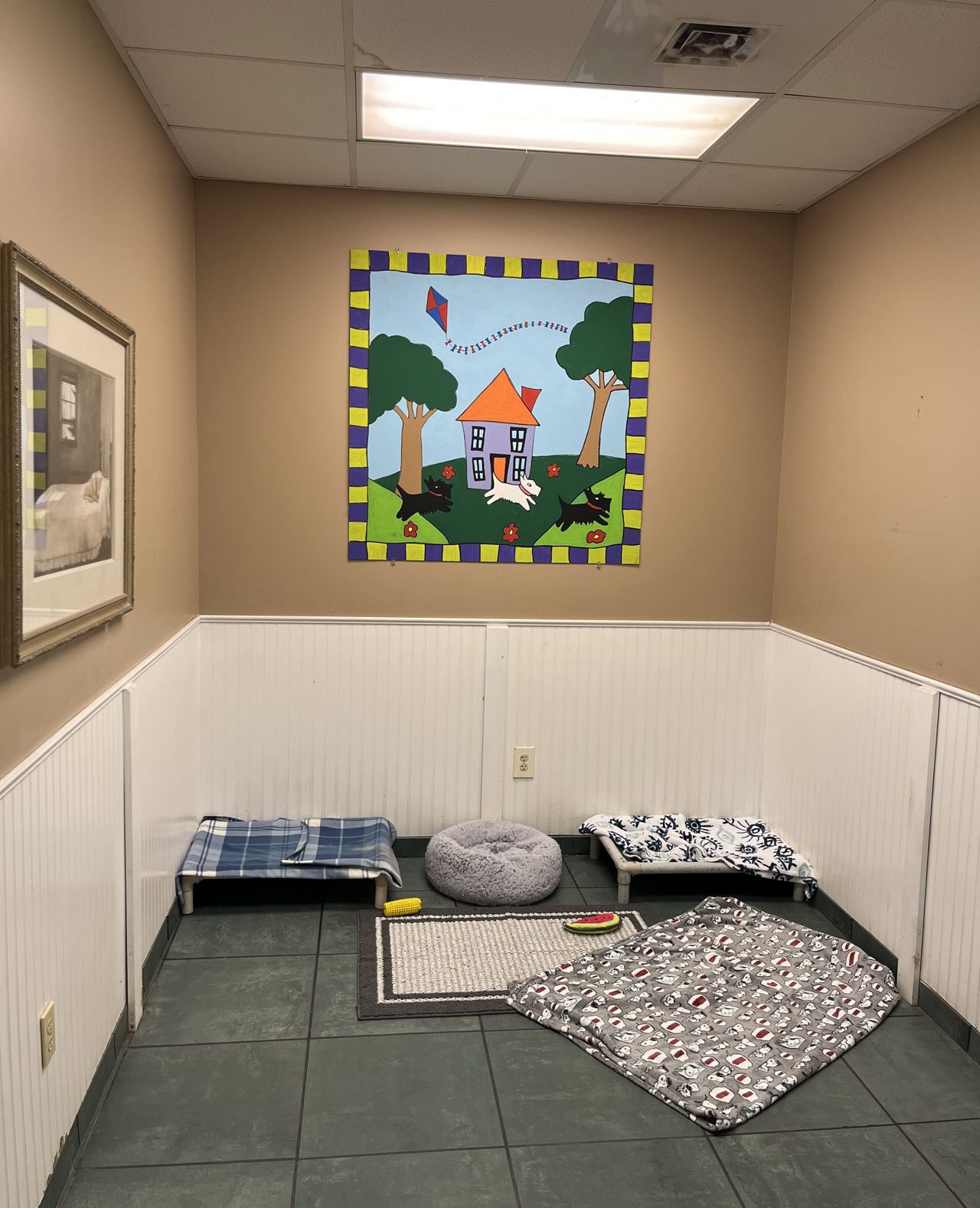 A clean, indoor dog boarding or daycare room featuring several cozy dog beds and a colorful, dog-themed quilt hanging on the wall