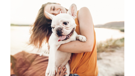 Smiling woman holding a happy white French Bulldog outdoors by a lake