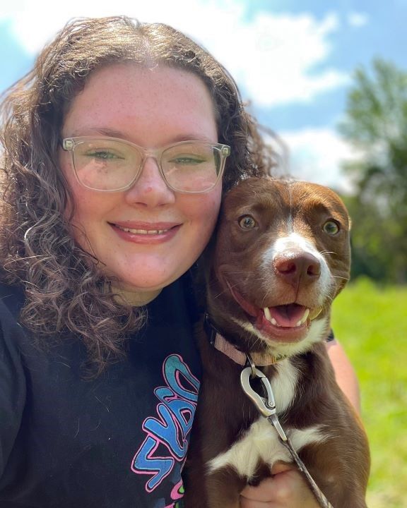 Brianna -Animal Care Attendant taking a selfie cheek to cheek with a happy chocolate and white mixed-breed puppy outdoors
