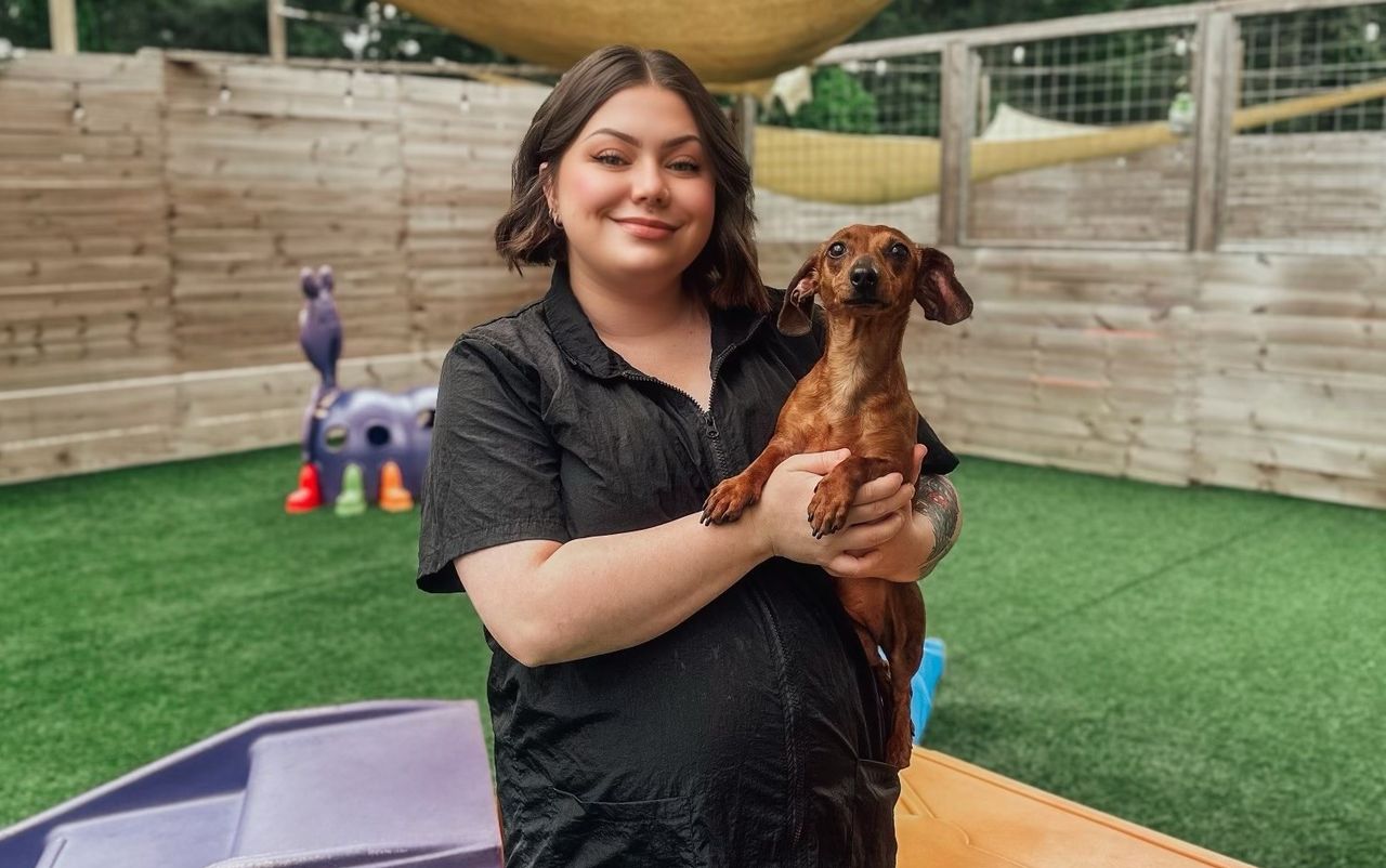 Texas Doghouse Groomer Stefani holds a small brown Dachshund while standing in an outdoor dog play area with green artificial turf
