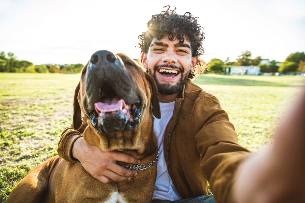 A happy young man with curly hair taking a wide-angle selfie in a grassy park, smiling broadly with his arm around a large brown dog with its mouth open