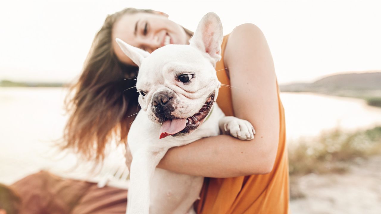 A smiling young woman holding a happy white French bulldog in her arms outdoors near the water