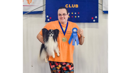 Trainer Jane holding a small black and white Papillon dog, displaying a gold medal and a blue competition award ribbon in front of a club backdrop