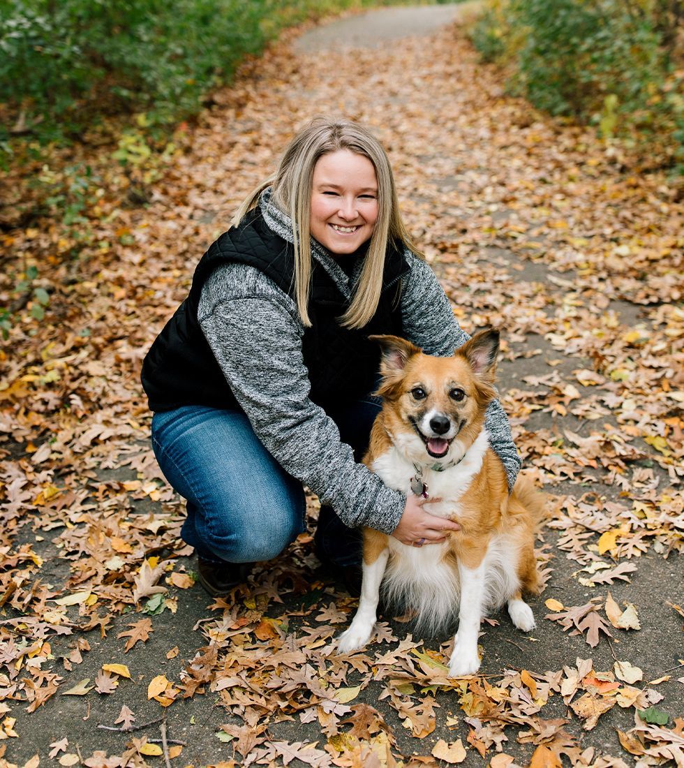 Paws and Pals Kasey -General Manager next to her medium-sized brown and white dog on a park path covered in autumn leavesSkylar