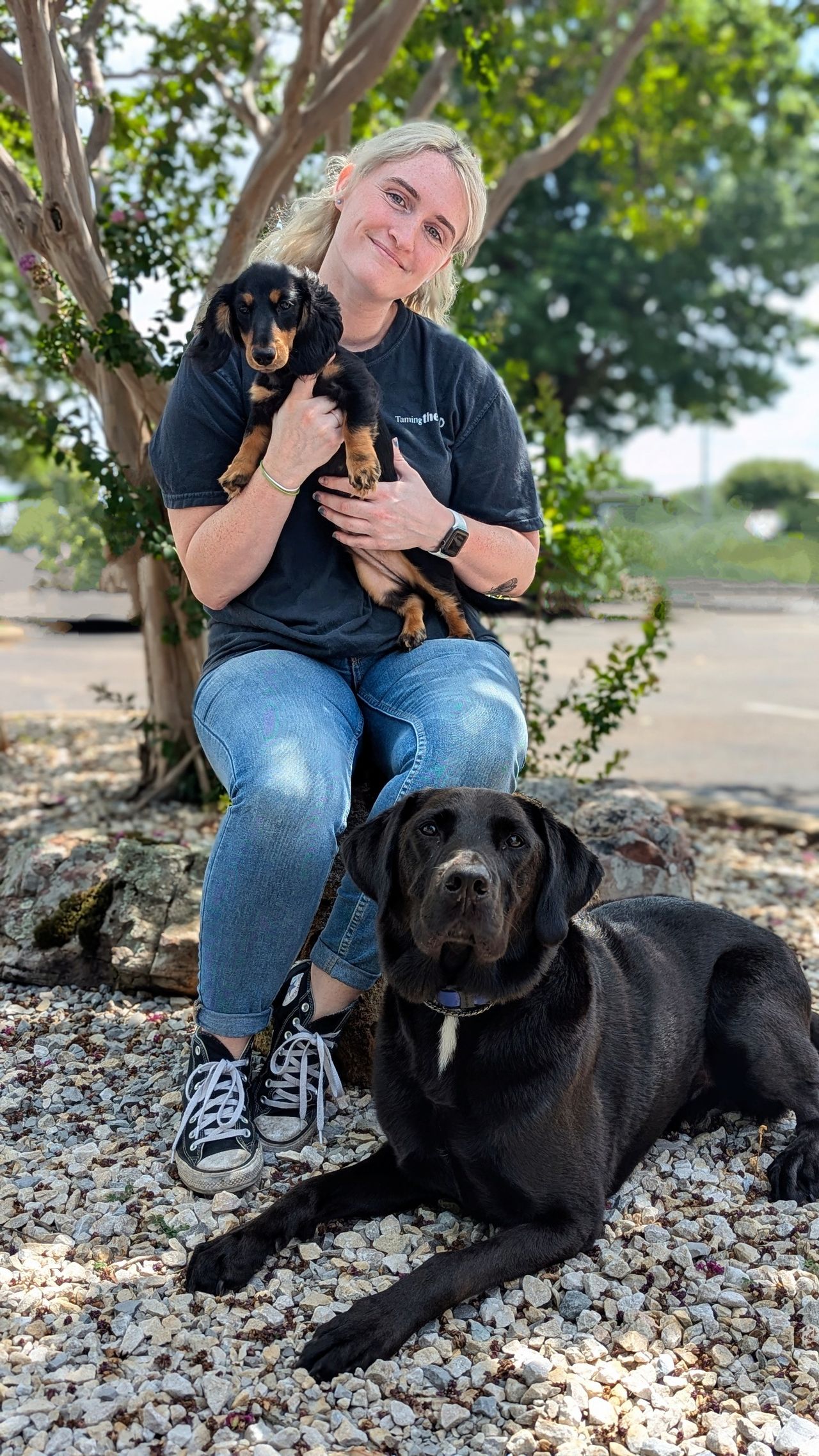 holding a small Dachshund puppy Ashley -Head Trainer sitting outdoors with a black Labrador mix resting at her feet