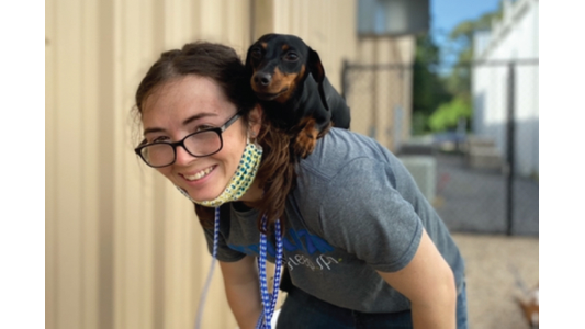 Smiling pet care staff member carrying a small black and tan Dachshund perched playfully on her back outdoors.