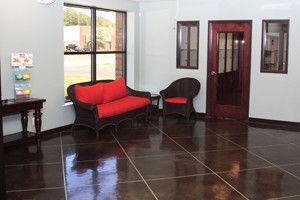 A waiting room with a dark floor, red cushioned wicker furniture, a door with glass panels, a window, and a pamphlet stand on a small table.