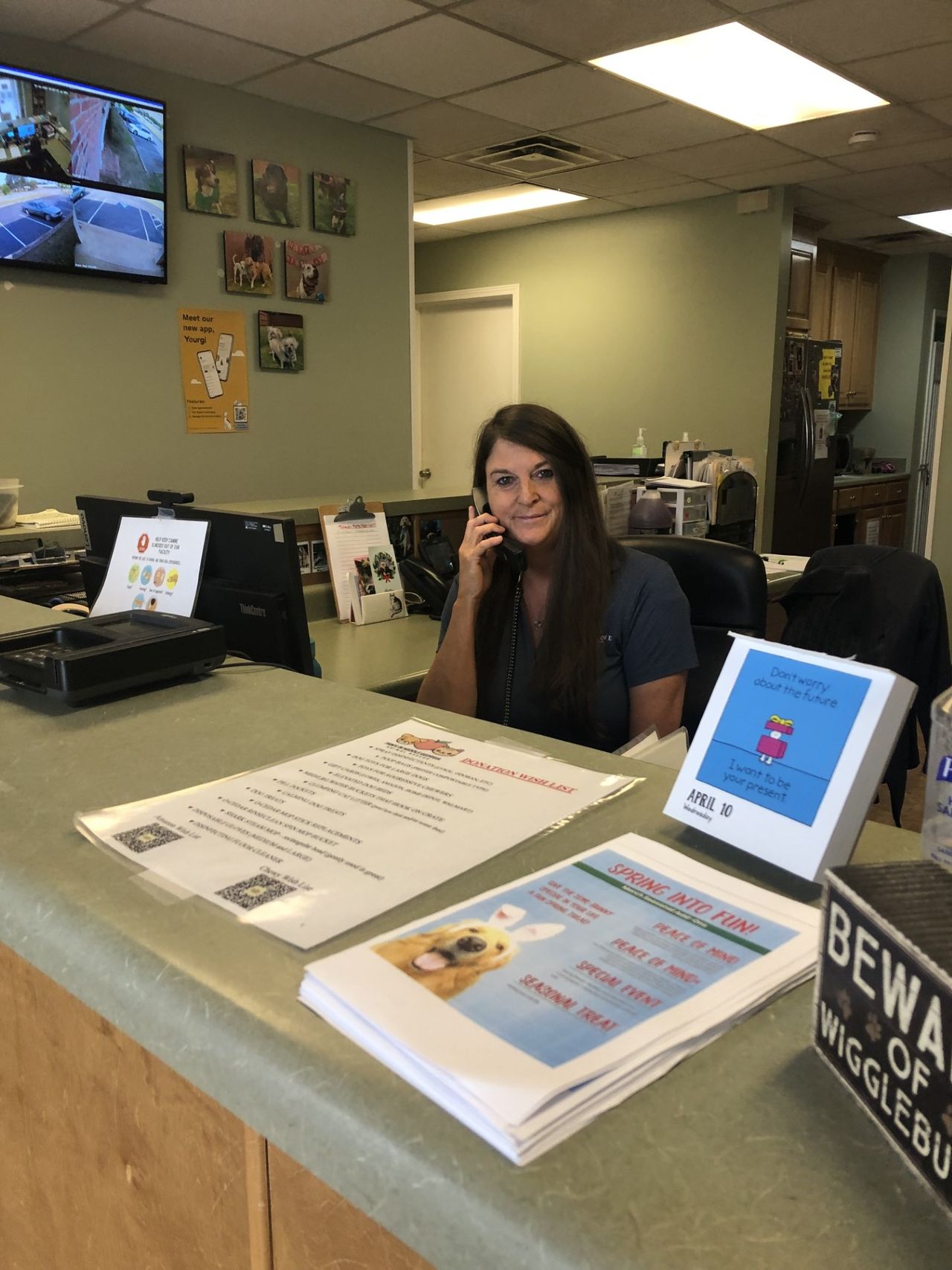 Donna Hall -Customer Service Representative behind a reception desk, holding a phone to her ear, with stacks of informational flyers and a "Beware of Wigglebutts" sign visible on the counter in the foreground.