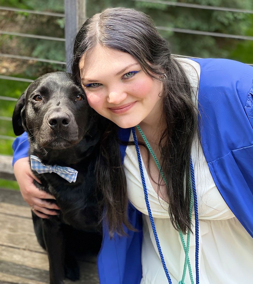 Lauren -Groomer hugging a black Labrador wearing a light blue gingham bow tie on a wooden deck