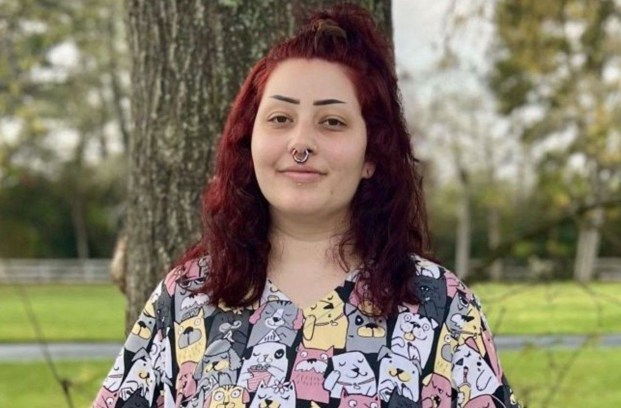 Texas Doghouse pack member Mikayla stands outdoors next to a tree trunk, wearing a top patterned with various cartoon dogs
