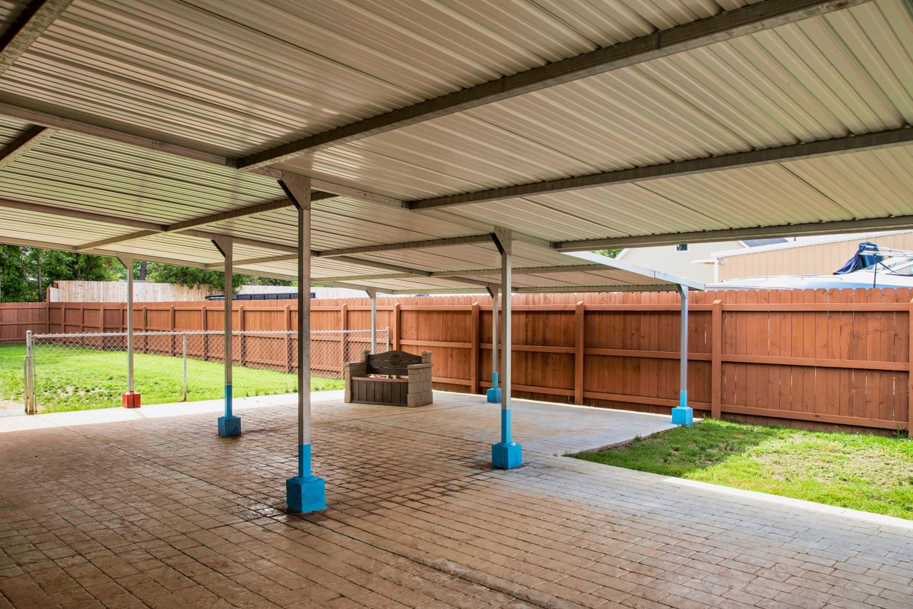 A large outdoor covered pavilion with a corrugated metal roof supported by metal poles with bright blue bases, shading a paved patio area. In the background, a small wooden bench sits under the shelter, next to a grassy yard enclosed by a tall brown wooden fence