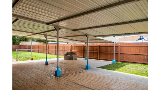 Endless Love Pet Palace covered pavilion with a corrugated metal roof supported by metal poles with bright blue bases, shading a paved patio area. In the background, a small wooden bench sits under the shelter, next to a grassy yard enclosed by a tall brown wooden fence